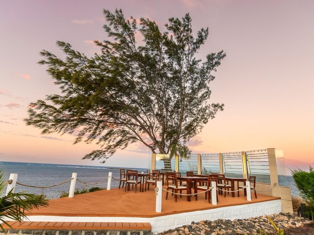 A breezy seaside deck with wooden tables and chairs under a tree