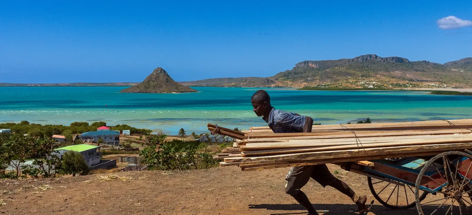 A man pulls a cart loaded with wood along the shore